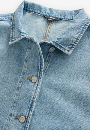 Lichtblauwe denimhemd met een puntkraag, voorzien van zilveren knopen en zichtbare stikseldetails. De stof is textuurrijk en iets vervaagd.
