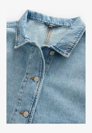 Camisa de mezclilla de color azul claro con cuello puntiagudo, que presenta botones plateados y detalles de costura visibles. La tela es texturada y ligeramente desgastada.