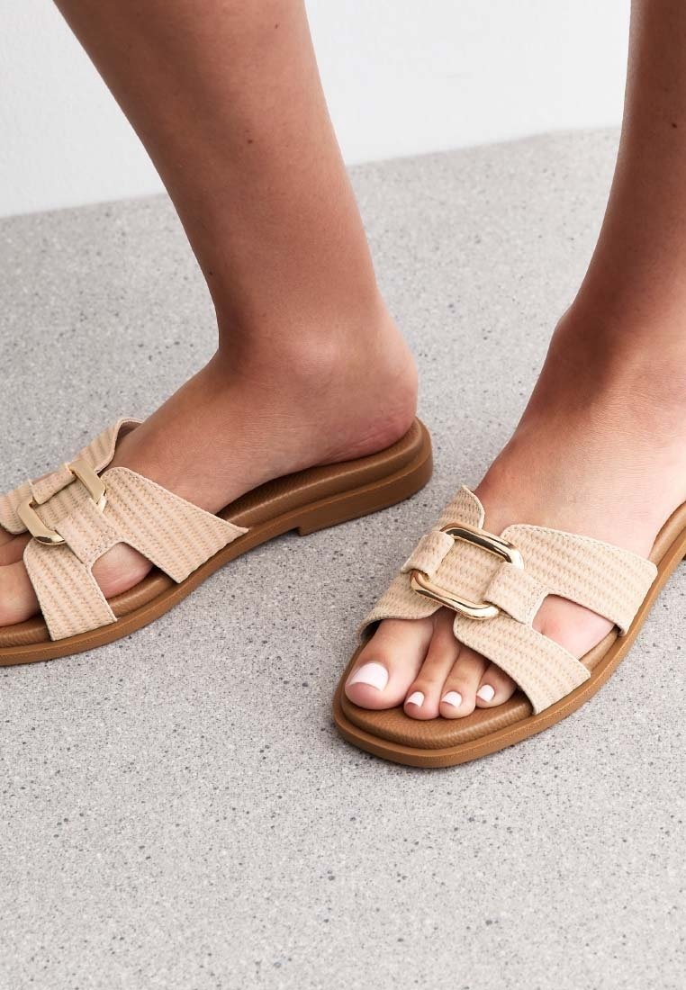 Des pieds portant des sandales beige à enfiler avec de larges sangles texturées et des boucles rectangulaires dorées sur un sol gris moucheté.