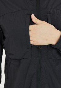 Hånd glider ind i brystlommen på en sort zip-jakke med elastisk manchet på ærmet.