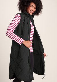 Woman smiling while wearing a long black quilted vest over a white and pink striped long-sleeve shirt, holding the vest open.
