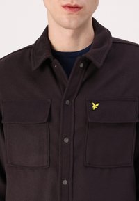 Veste noire à boutons avec col, deux poches poitrine et un petit logo d'aigle jaune sur la poche gauche. Tissu texturé.