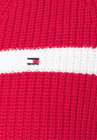 Rood gebreide stof met een witte horizontale streep en een klein geborduurd logo met blauwe, witte en rode details.