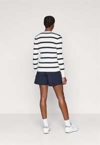 Hvit og marineblå stripete ribbestrikket genser, rund hals, tettsittende design. Kledd med marineblå shorts, båret med hvite joggesko og ankelstrømper.