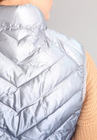 Padded silver väst med en chevron-quiltad mönster, designad med en slät, glänsande textur och en hög krage för extra värme.