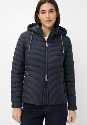 Frau trägt eine schwarze gesteppte Kapuzenjacke mit einem doppelten Frontreißverschluss und gestreiften Kordeln, kombiniert mit dunklen Jeans vor weißem Hintergrund.