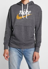 Grå huvtröja med en ficka fram, vita snoddar och en stor gul Nike-logotyp på framsidan. Mjukt och texturerat tyg.