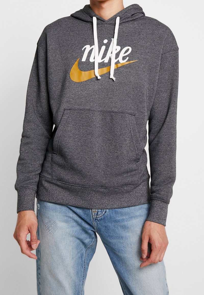 Grå huvtröja med en ficka fram, vita snoddar och en stor gul Nike-logotyp på framsidan. Mjukt och texturerat tyg.