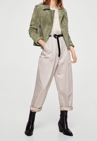 Grüne Bikerjacke aus Veloursleder, cremefarbenes Oberteil, beige plissierte Hose mit Bündchen, schwarze Taillenbetonung durch Gürtel und schwarze Stiefeletten mit Blockabsatz.