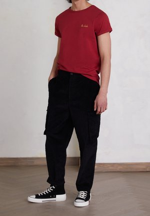 Mann trägt ein rotes T-Shirt mit hochgekrempelten Ärmeln und der Aufschrift "the dude", schwarze Cord-Cargohosen und schwarze High-Top-Sneaker, steht drinnen.