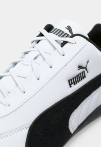 Hvide sneakers lavet af læder og syntetiske materialer; har sorte accenter, strukturerede paneler og et logo på siden med snørebånd.