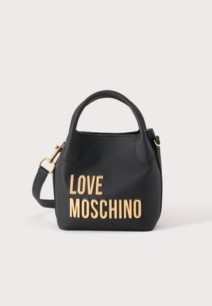 Čierna kabelka Moschino so zlatými nápismi, krátkymi rúčkami a odnímateľným ramenným popruhom na jednofarebnom svetlom pozadí.