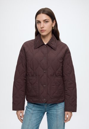 Kvinde iført en brun quiltet jakke med store frontlommer og en fløjlskrave, kombineret med lyseblå jeans, stående mod en hvid baggrund.