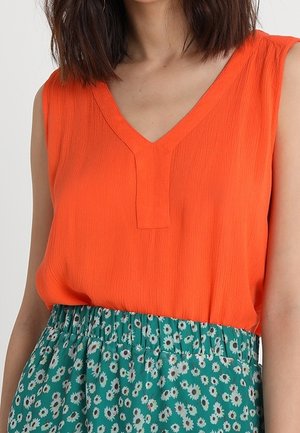 Ärmelloses, leuchtend oranges, V-förmiges Oberteil kombiniert mit einem hoch taillierten grünen Rock mit kleinem weißem Blumenmuster.