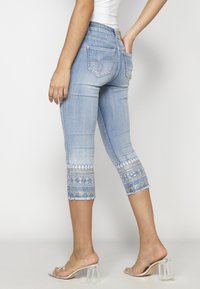Jean cropped bleu clair avec une broderie blanche et bleue complexe aux poignets, portés avec des sandales à talons transparents et un haut blanc.