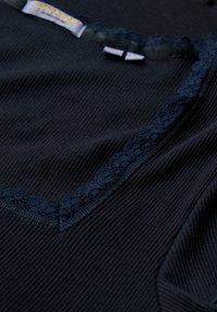 Sort ribbet stof med en blondekant langs halsen, der har en glat tekstur og en subtil glans. Inkluderer et mærke med brandingdetaljer.