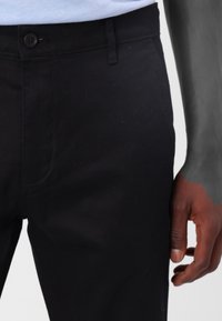 Pantalon noir en coton avec une texture lisse, coupe droite et une fermeture à bouton à la taille, avec des poches latérales visibles.