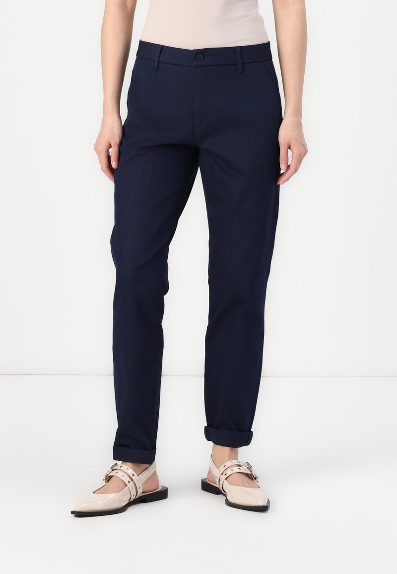 Femme portant un pantalon slim bleu marine avec des ourlets retroussés et des chaussures plates beige clair avec des brides et des clous.