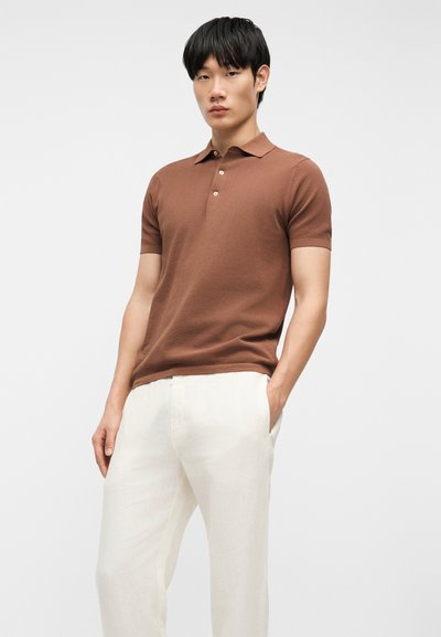 Jeune homme portant un polo marron à manches courtes et un pantalon blanc, debout avec une main dans la poche devant un fond uni.