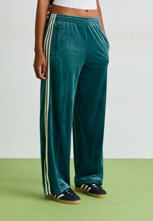 Person iført turkisblå velour træningsbukser med hvide striber på siderne og sorte sneakers med hvide striber, stående på grøn gulv.