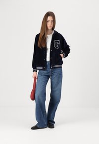 Mornarica samet varsity jakna s bijelim prugama i oznakom "G", nosi se preko bijele majice, uparena s širokim plavoijeans hlačama i crnim cipelama.