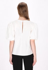 Hvid bluse med puffede ærmer, detalje med nøglehul bagpå og glat tekstur. Afsluttet med en rund halsudskæring og en let buet kant.
