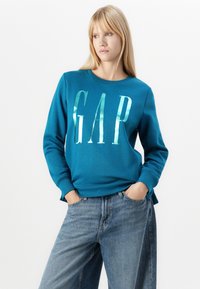 Turkoosi collegepaita, jossa on metallinvärinen "GAP"-logo, pyöreä kaula-aukko, ribattu hihansuu ja rento leikkaus, yhdistettynä vaaleansinisiin leveälahkeisiin farkkuihin.