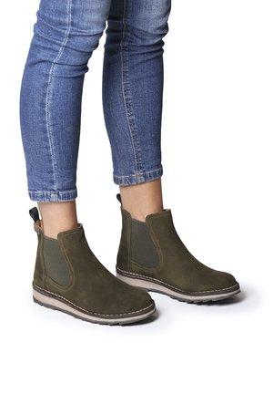 Bottines en suède vert olive avec panneaux latéraux élastiques et tirant. Semelle en caoutchouc avec coutures contrastées et petit talon. Jeans visibles au-dessus des bottines.
