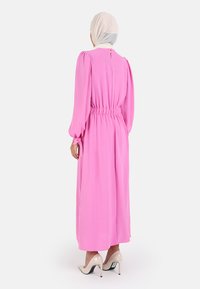 Lang pink kjole med puffærmer og indsnævret talje, med en åbning i nakken. Bæres med beige højhælede sko.