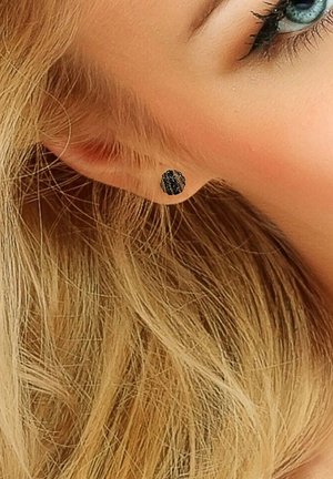 Boucle d'oreille clou noire texturée, en forme ronde, présentant une surface texturée qui semble brillante, placée sur un lobe d'oreille perforé.