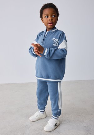 Młody chłopiec ubrany w niebiesko-biały dres i białe trampki, stoi na szarej podłodze, patrząc na bok z złożonymi rękami.