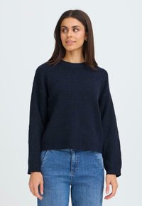 Maglione a coste in blu navy, con maniche lunghe e colletto a girocollo, abbinato a jeans in denim. La texture sembra morbida e spessa.