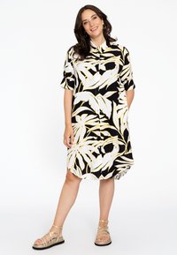 Schwarz kurzärmeliges Kleid mit weißen und gelben Blattmustern, mit Kragen, Knopfverschluss und abgerundetem Saum. Getragen mit goldenen Sandalen.