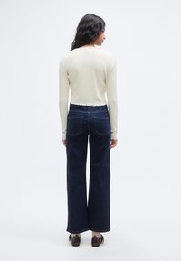 Ανοιχτό κρεμ μακρυμάνικη μπλούζα με υφασμένο σχέδιο και κυματιστό τελείωμα, συνδυασμένη με σκούρα μπλε τζιν παντελόνι φαρδιάς γραμμής, εμφανιζόμενα από πίσω.