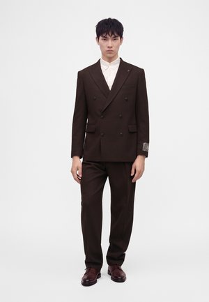 Jovem vestido com fato de corte duplo castanho escuro, camisa branca e sapatos castanhos, posando contra um fundo claro e liso.