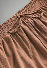 Braunes, gefälteltes Gewebe mit einem Smock-Taillengürtel und einer Schleifendetail an der Vorderseite. Die Textur ist zerknittert, was ein entspanntes Aussehen erzeugt.