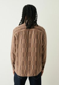 Brun mønstret skjorte med en tekstureret stof, prydet med et ternet design og lange ærmer, set fra bagsiden.