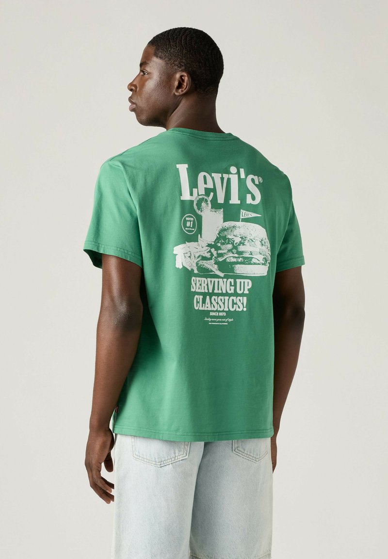Mann trägt grünes Levi's T-Shirt mit weißem Motiv eines Burgers, Pommes und Getränks sowie dem Schriftzug „Serving Up Classics!“ auf dem Rücken.