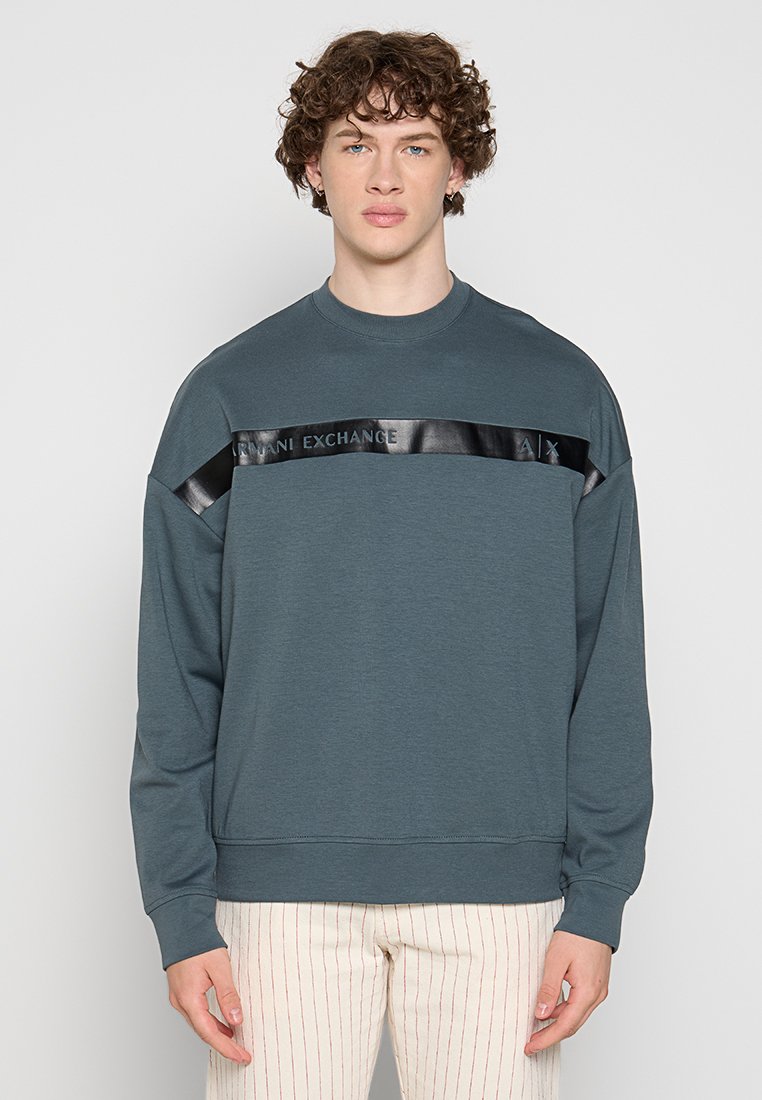 Junger Mann mit lockigem Haar, der einen dunkelblauen Pullover mit einem glänzenden schwarzen "Armani Exchange"-Streifen über der Brust und einer hellen Hose mit feinen vertikalen Streifen trägt.