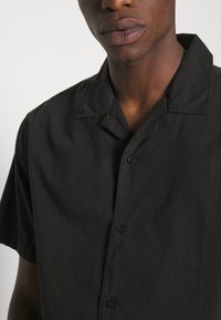 Černá košile s krátkým rukávem a klasickým límcem, vyrobená z hladké tkaniny, s předními knoflíky a volným střihem.