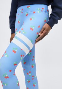 Personne portant des leggings bleus avec de petits imprimés fraises rouges et fleurs blanches et deux bandes blanches horizontales sur la cuisse.