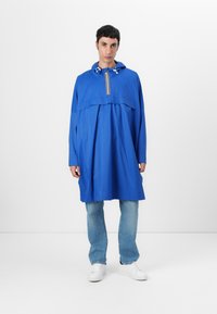 Blå huva poncho med dragkedja fram, som har en lös passform, långa ärmar och snörningsdetaljer runt huvan. Bärs med ljusa denimjeans.