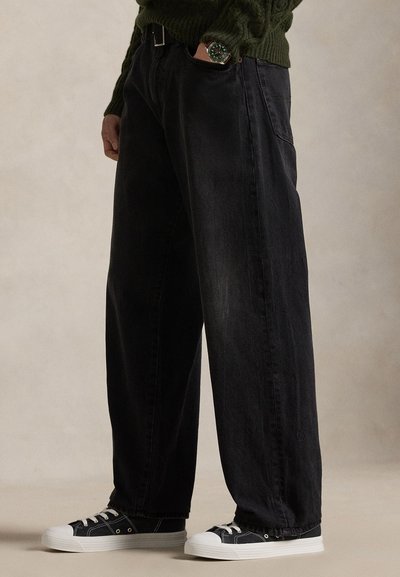 Person som har på seg vide svarte jeans, svart-hvite joggesko og en grønn genser, står med en hånd i lomma mot en beige bakgrunn.
