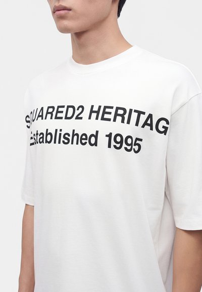 Άνδρας που φοράει λευκό t-shirt με μαύρο κείμενο που γράφει "SQUARED2 HERITAG Established 1995" τυπωμένο στο στήθος.