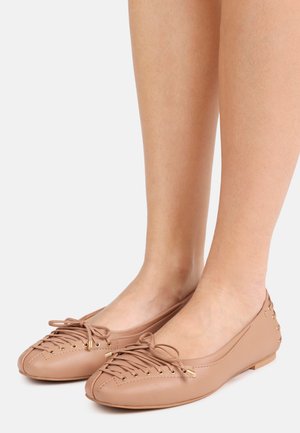 Gambe nude che indossano ballerine in pelle marrone chiaro con lacci decorativi e piccoli borchietti metallici sul tallone.