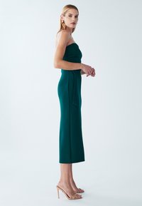 Vestito midi color turchese senza spalline, con silhouette aderente e tessuto strutturato. Abbinato a sandali con tacco color nudo, che esaltano una figura snella.