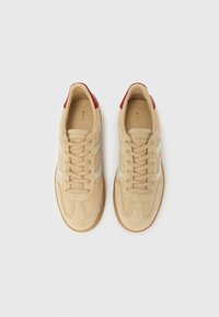 Beige ruskindssneakers med hvide sidelisser, beige snørebånd, gummisåler og røde læderhæl-accenter, set oppefra på en hvid baggrund.