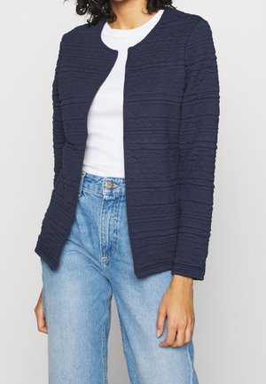 Personne portant une veste bleu marine texturée, ouverte sur le devant, sur un haut blanc avec un jean taille haute bleu clair.