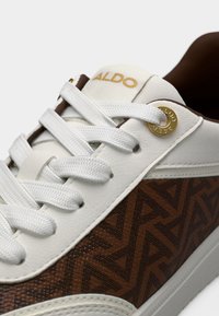 Baskets blanches et marron avec motif géométrique, lacets blancs, œillet en métal doré et logo "ALDO" sur la languette.