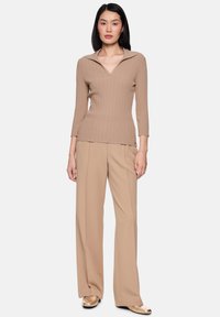 Frau trägt beigefarbenen, gerippten V-Ausschnitt-Pullover mit dreiviertellangen Ärmeln, dazu passende weite Hosen und metallisch goldene Slipper, steht vor weißem Hintergrund.